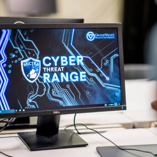 A computer monitor displays a digital interface with the text "CYBER THREAT RANGE" in large letters, along with the logos of MiCTRA and GVSU. The background features a blue and black circuit board design, and the monitor sits on a a white desk.